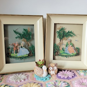 May include: Two vintage framed pictures with a cream colored mat and gold frame. The pictures are of a couple in a garden setting and a woman sitting under a tree. The pictures are sitting on a crocheted blanket with two small owl and swan figurines.