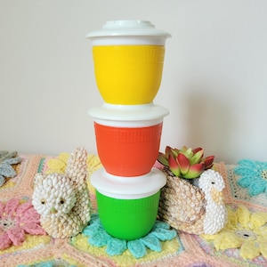 May include: Three stackable plastic containers with lids. The top container is yellow, the middle container is orange, and the bottom container is green. The containers are on a crocheted surface.