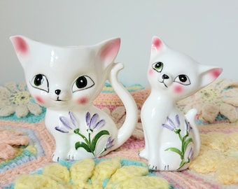 May include: Two white ceramic cat figurines with purple flowers painted on them. The cat on the left has a curled tail and the cat on the right has its ears perked up.