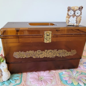 May include: A vintage amber plastic storage box with a floral design and a gold latch. The box is sitting on a crocheted blanket with a crocheted owl figurine on top.