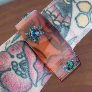 May include: A brown and amber plastic bangle bracelet with blue and green rhinestones. The bracelet has two clusters of rhinestones.