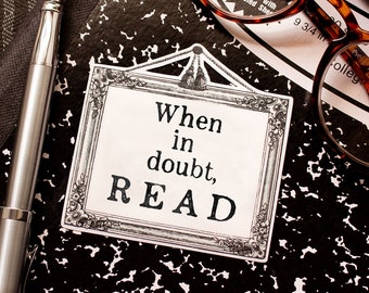 May include: A black and white framed sign with the text "When in doubt, READ" on a black and white speckled background. A pair of brown tortoiseshell glasses and a silver pen are also in the image.