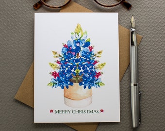 Può includere: Un biglietto di Natale con un'illustrazione ad acquerello di un arrangiamento di fiori di campanula blu a forma di albero di Natale in un vaso marrone. Il biglietto ha il testo "Merry Christmas" stampato sotto l'illustrazione.