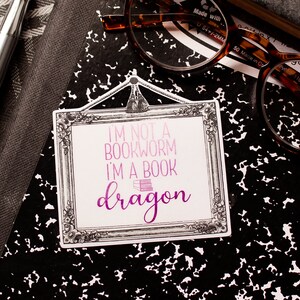 May include: A sticker with the text "I'M NOT A BOOKWORM I'M A BOOK dragon" in pink and purple, framed in a decorative border. The sticker rests on a black and white composition notebook, with a pen and glasses nearby.