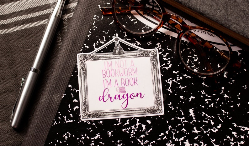 May include: A sticker with the text "I'M NOT A BOOKWORM I'M A BOOK dragon" in pink and purple, framed in a decorative border. The sticker rests on a black and white composition notebook, with a pen and glasses nearby.