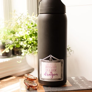 May include: A black water bottle with a sticker that reads "I'm not a bookworm I'm a book dragon." The bottle is sitting on a stack of books, with a pair of glasses and a pen nearby. The bottle has a black lid.