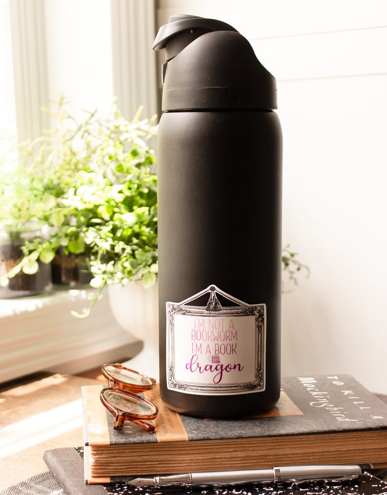 May include: A black water bottle with a sticker that reads "I'm not a bookworm I'm a book dragon." The bottle is sitting on a stack of books, with a pair of glasses and a pen nearby. The bottle has a black lid.