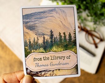Puede incluir: Una pegatina rectangular con una ilustración de paisaje vintage que presenta una montaña, árboles y un cielo nublado. La pegatina tiene un borde blanco y el texto "from the library of Thomas Gunderson".
