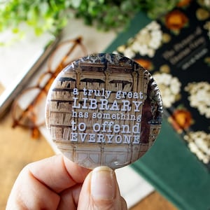 May include: A round button with the text "a truly great LIBRARY has something to offend EVERYONE" over a background of bookshelves. The button is held in a hand, with glasses and a book visible in the background.
