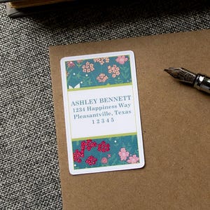 May include: A white return address label with a floral design in shades of pink, green, and white. The label has the text "ASHLEY BENNETT 1234 Happiness Way Pleasantville, Texas 12345".