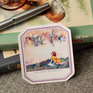 May include: A bookplate featuring a whimsical illustration of fairies flying above a young girl sitting in a field. The text "THIS BOOK BELONGS TO" is printed at the bottom of the bookplate.
