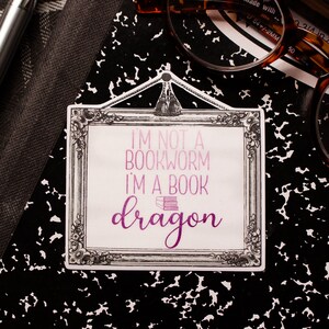 May include: A rectangular sticker with a decorative frame and the text "I'M NOT A BOOKWORM I'M A BOOK dragon" in shades of purple. The sticker is on a black and white speckled surface, with a pen and glasses in the background.