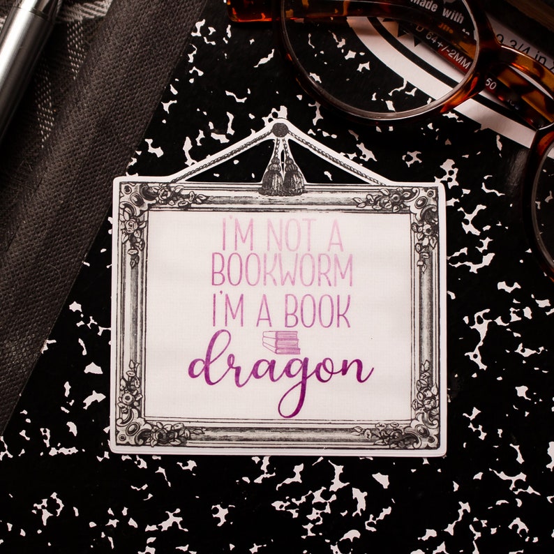 May include: A rectangular sticker with a decorative frame and the text "I'M NOT A BOOKWORM I'M A BOOK dragon" in shades of purple. The sticker is on a black and white speckled surface, with a pen and glasses in the background.