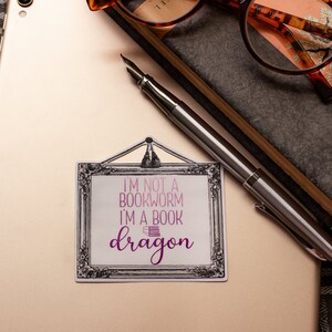 May include: A sticker with the text "I'M NOT A BOOKWORM I'M A BOOK dragon" in pink and purple, framed in a decorative black and white border. A silver fountain pen rests on a light-colored surface, near a pair of glasses and a notebook.