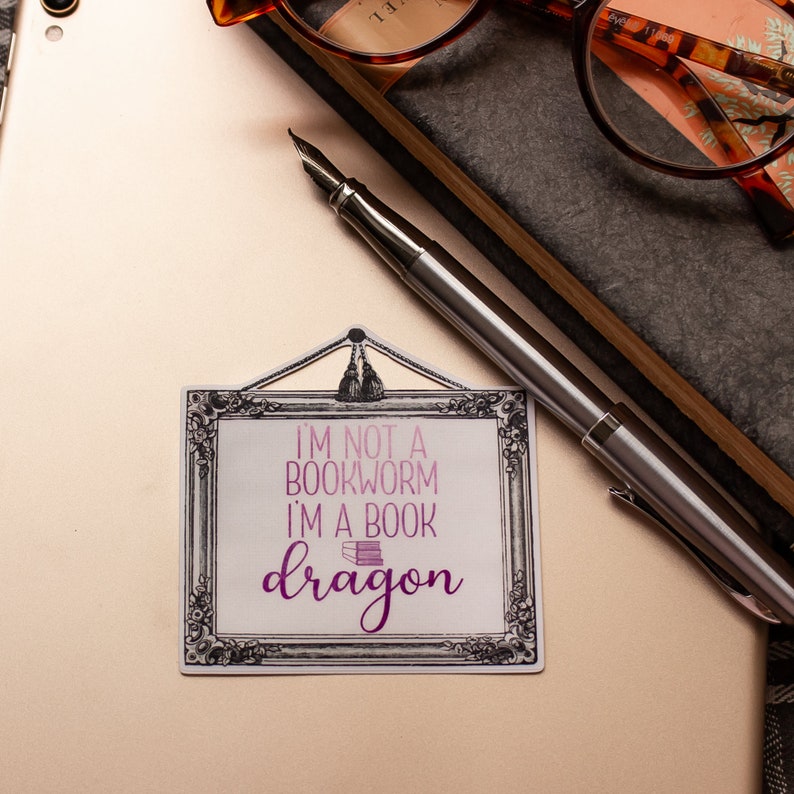 May include: A sticker with the text "I'M NOT A BOOKWORM I'M A BOOK dragon" in pink and purple, framed in a decorative black and white border. A silver fountain pen rests on a light-colored surface, near a pair of glasses and a notebook.