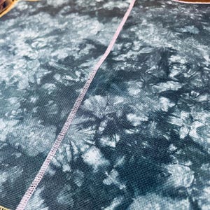 Peut inclure: Gros plan sur un tissu avec un motif tie-dye dans les tons de bleu sarcelle et de blanc. Une couture rose clair traverse l'image en diagonale. Le tissu a un aspect texturé.