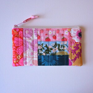 May include: A rectangular, quilted zipper pouch with a variety of floral and fruit patterns. The pouch features a pink zipper pull and is made of fabric with pink, red, blue, and yellow floral and strawberry designs.