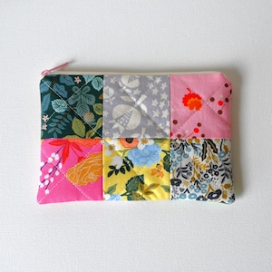 May include: A rectangular quilted zipper pouch featuring a patchwork design of six distinct fabric squares. The patterns include dark green foliage, grey pomegranates, pink floral motifs, bright pink flowers, yellow blossoms, and a dense blue floral print.