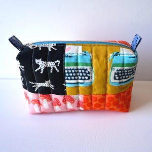 May include: A patchwork zippered pouch with a variety of fabric patterns. The pouch features panels of black fabric with white cat silhouettes, pink fabric with red mushrooms, and fabric with vintage typewriter illustrations. The zipper is blue.
