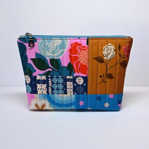 May include: A quilted cosmetic bag with a patchwork design. The bag features a variety of floral and geometric patterns in pink, blue, orange, and brown. It has a blue zipper and is approximately 9 inches wide and 6 inches tall.