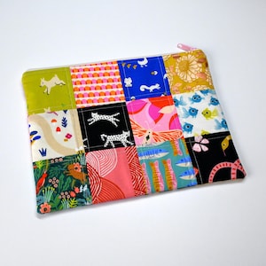May include: A patchwork zippered pouch with a variety of colorful fabric squares. The squares feature whimsical designs including running cats, fish, and floral patterns. The zipper is pink, and the pouch is rectangular.