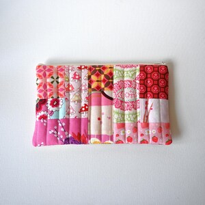 May include: A rectangular quilted zipper pouch featuring a patchwork design of various fabrics in shades of pink, red, and white. The patterns include mushrooms, strawberries, floral motifs, and geometric shapes, all stitched together with vertical quilting lines.