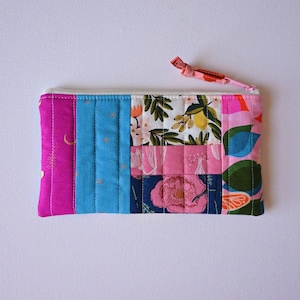 May include: A quilted pouch with a white zipper and a pink fabric pull. The pouch is made of various fabric panels in shades of pink, blue, and floral patterns. The pouch is rectangular and has vertical stitching.