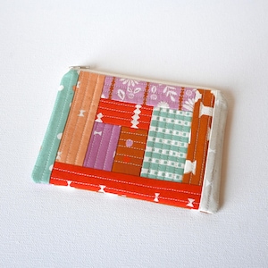 May include: A colorful, handmade zippered pouch with a patchwork design. The pouch features a variety of fabrics in shades of orange, pink, green, and white, stitched together in a geometric pattern. The zipper is silver.
