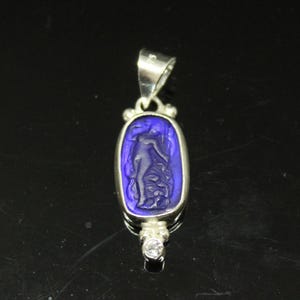 May include: A silver pendant with a rectangular, deep blue center featuring a carved figure. The pendant has a small, clear gemstone at the bottom and a silver loop at the top for a chain. The design is classic and elegant.