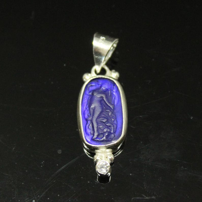 May include: A silver pendant with a rectangular, deep blue center featuring a carved figure. The pendant has a small, clear gemstone at the bottom and a silver loop at the top for a chain. The design is classic and elegant.