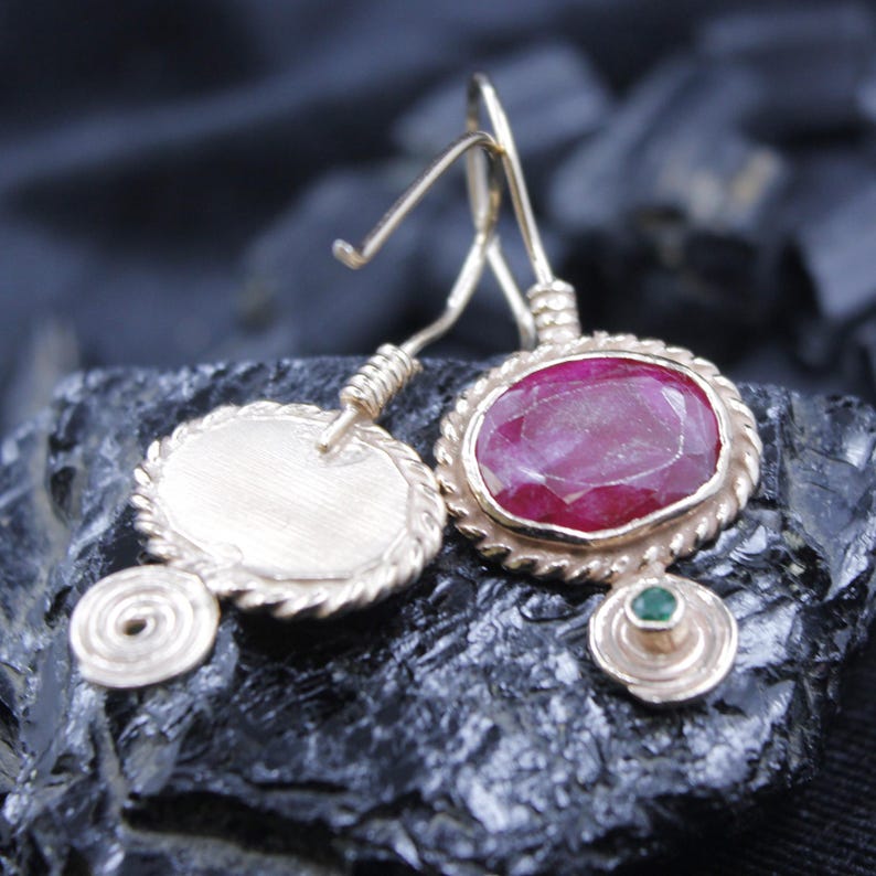 May include: Gold-toned earrings with a ruby-colored oval stone and a smaller green stone. The earrings have a textured, rope-like border around the stones and a spiral design. The earrings are displayed on a dark, textured surface.