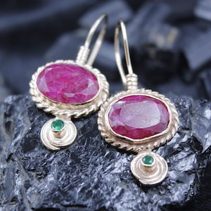 May include: A pair of gold-toned earrings featuring large, oval-cut ruby gemstones. Each earring has a twisted rope-like border around the ruby and a small, round green gemstone at the bottom. The earrings are displayed on a dark, textured surface.