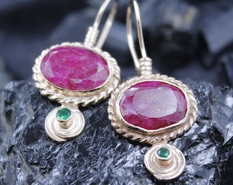 May include: A pair of gold-toned earrings featuring large, oval-cut ruby gemstones. Each earring has a twisted rope-like border around the ruby and a small, round green gemstone at the bottom. The earrings are displayed on a dark, textured surface.