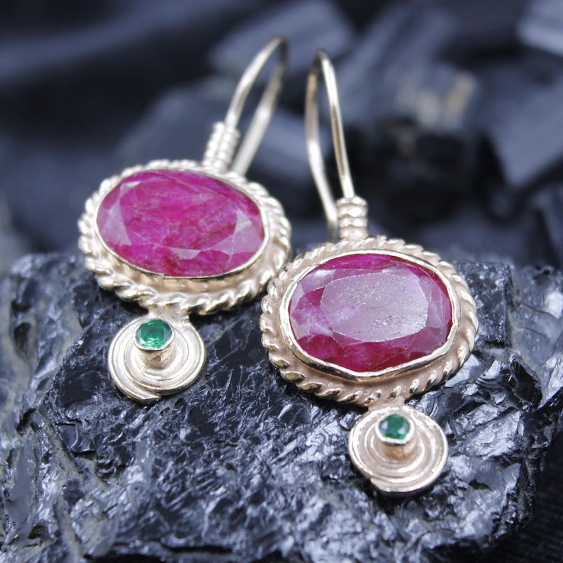 May include: A pair of gold-toned earrings featuring large, oval-cut ruby gemstones. Each earring has a twisted rope-like border around the ruby and a small, round green gemstone at the bottom. The earrings are displayed on a dark, textured surface.