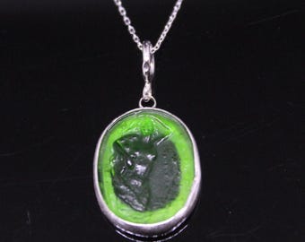 Puede incluir: Un collar de plata con un colgante ovalado. El colgante presenta una figura verde oscuro sobre un fondo verde brillante. El colgante está enmarcado por un borde plateado y cuelga de una delicada cadena plateada.