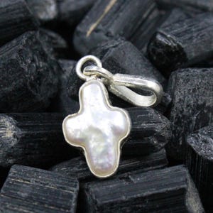 May include: A silver-toned cross pendant with a pearlescent, off-white center. The cross has a rounded shape and is set against a backdrop of dark, textured stones. The pendant includes a loop for a chain.