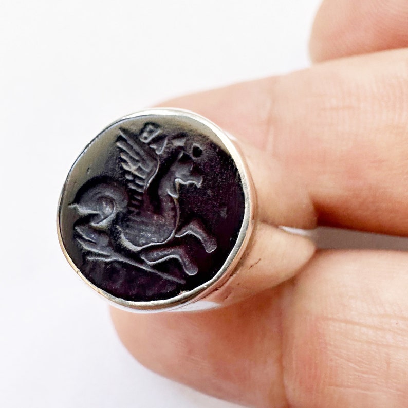 May include: A silver ring with a black onyx stone featuring a carved image of a winged horse.