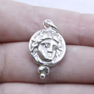 May include: Silver pendant featuring a detailed profile of a woman's face with a crown of leaves. The pendant has a round shape and a small, three-bead chain attached to the bottom.