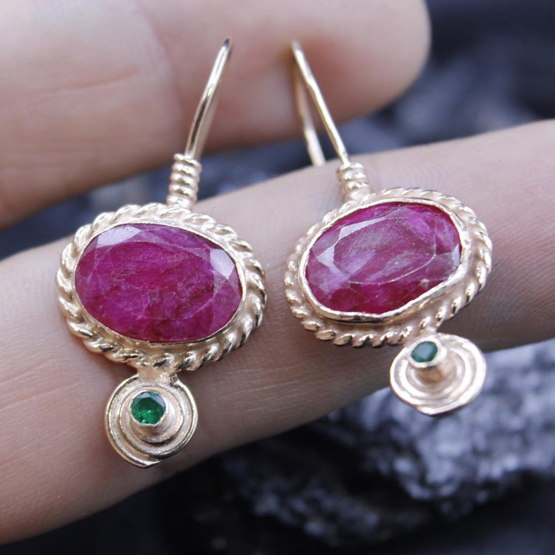 May include: A pair of gold-toned earrings featuring oval-shaped, deep red gemstones. Each earring has a twisted rope-like border around the main stone and a small, round green gemstone at the bottom. The earrings have a hook closure.
