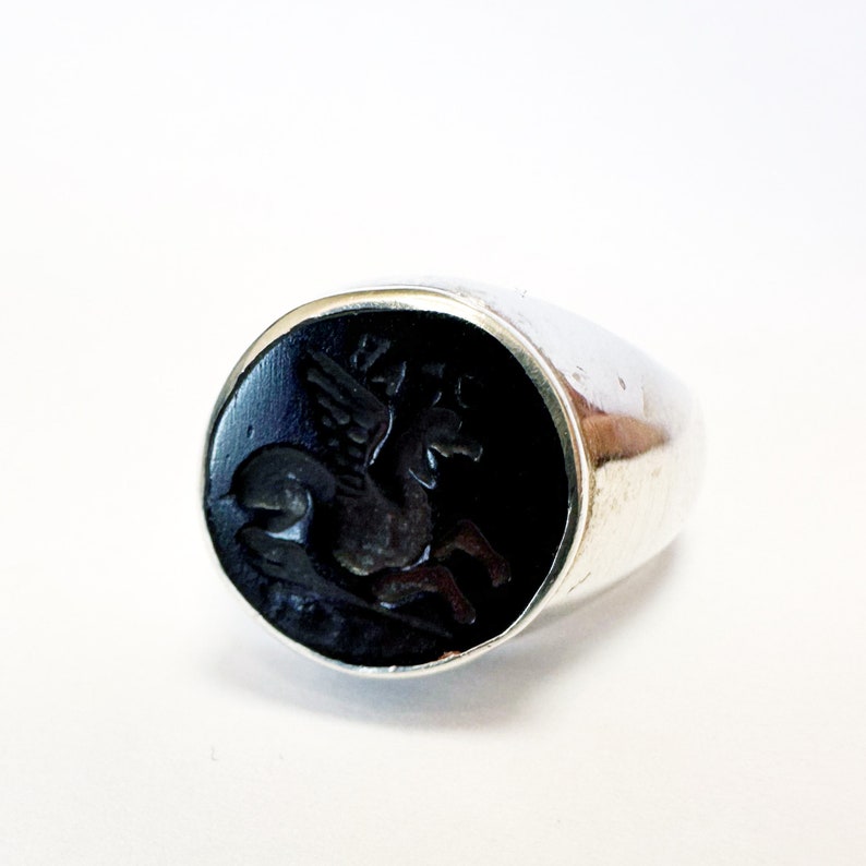 May include: A silver ring with a black onyx stone featuring a carved image of a winged horse.
