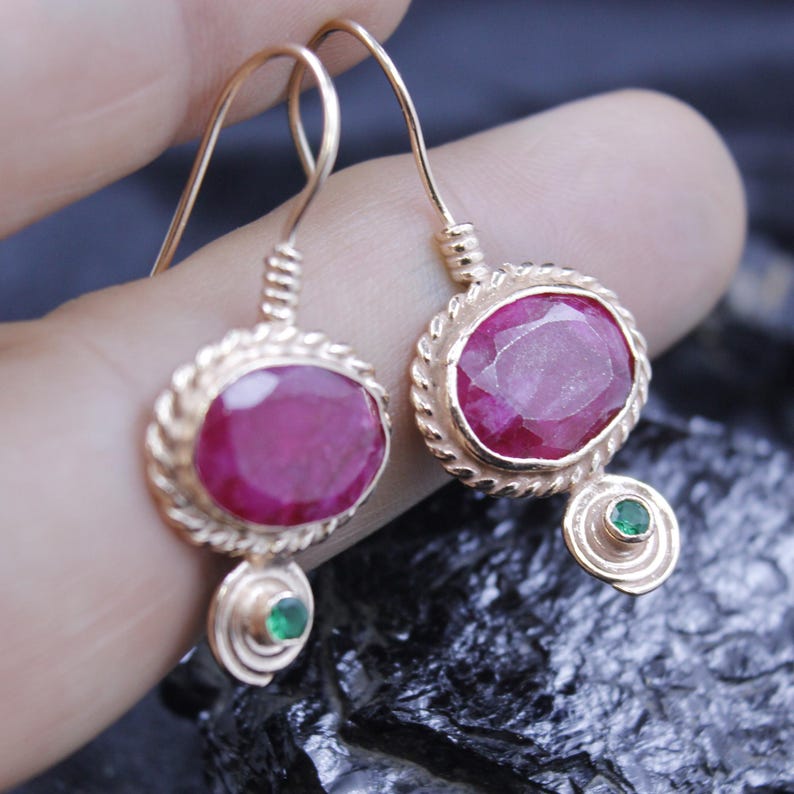 May include: Gold-toned drop earrings featuring large, oval-cut ruby gemstones. Each earring has a smaller, round green gemstone accent. The earrings have a textured, rope-like border around the ruby and green gemstones.