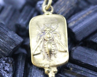 May include: A gold-colored pendant with a rectangular shape and a detailed insect design. The pendant has a loop at the top for a chain and a decorative bottom. The insect is centered on the pendant.