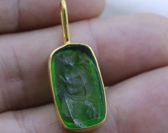 Puede incluir: Un colgante de vidrio verde con marco dorado y una talla detallada de una figura. El colgante está en una cadena de oro.