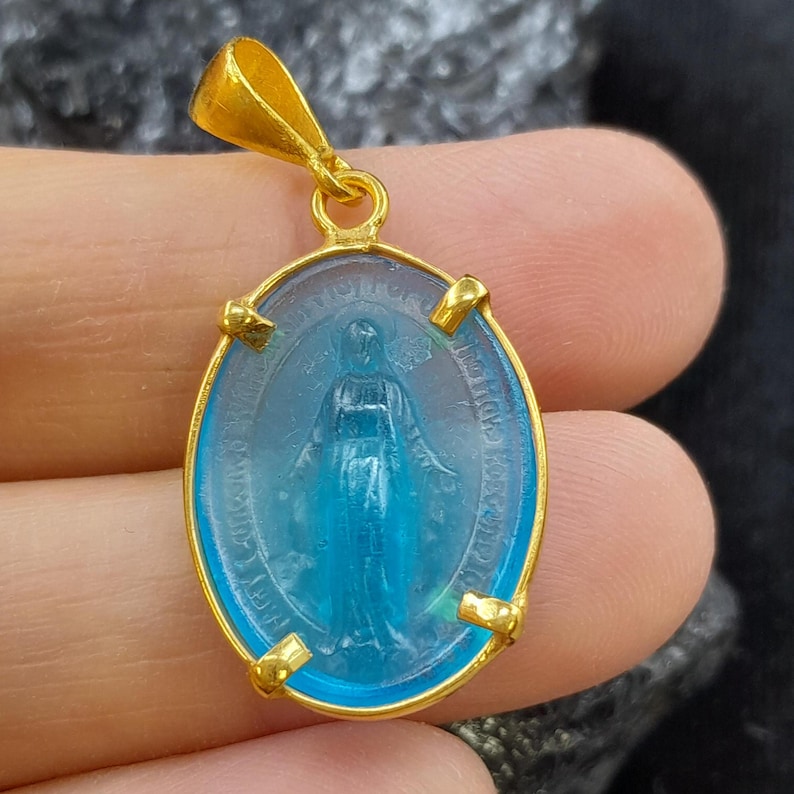 Puede incluir: Colgante dorado con un &oacute;valo de vidrio azul con una imagen en relieve de la Virgen Mar&iacute;a. El colgante tiene un aro dorado y cuatro acentos dorados alrededor del &oacute;valo.