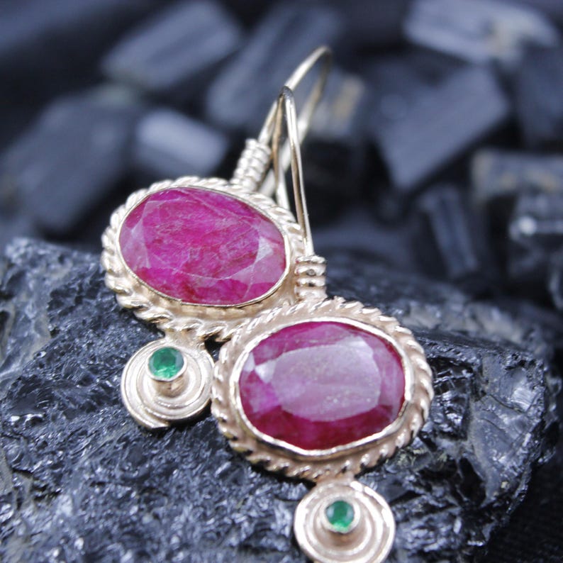 May include: Gold-toned earrings featuring large, oval-cut ruby gemstones. Each earring has a rope-like border around the ruby and a small, round emerald accent. The earrings are displayed against a dark, textured background.
