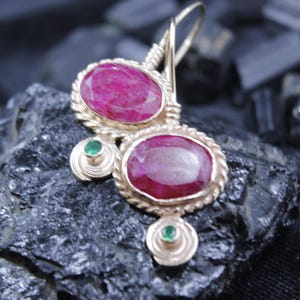 May include: Gold-toned earrings featuring two large, oval-shaped, ruby-colored gemstones. The earrings have a twisted rope design around the rubies and small green gemstones. The earrings are displayed on a dark, textured surface.