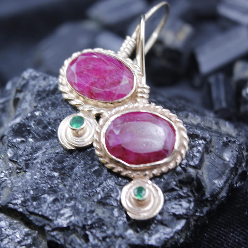 May include: Gold-toned earrings featuring two large, oval-shaped, ruby-colored gemstones. The earrings have a twisted rope design around the rubies and small green gemstones. The earrings are displayed on a dark, textured surface.