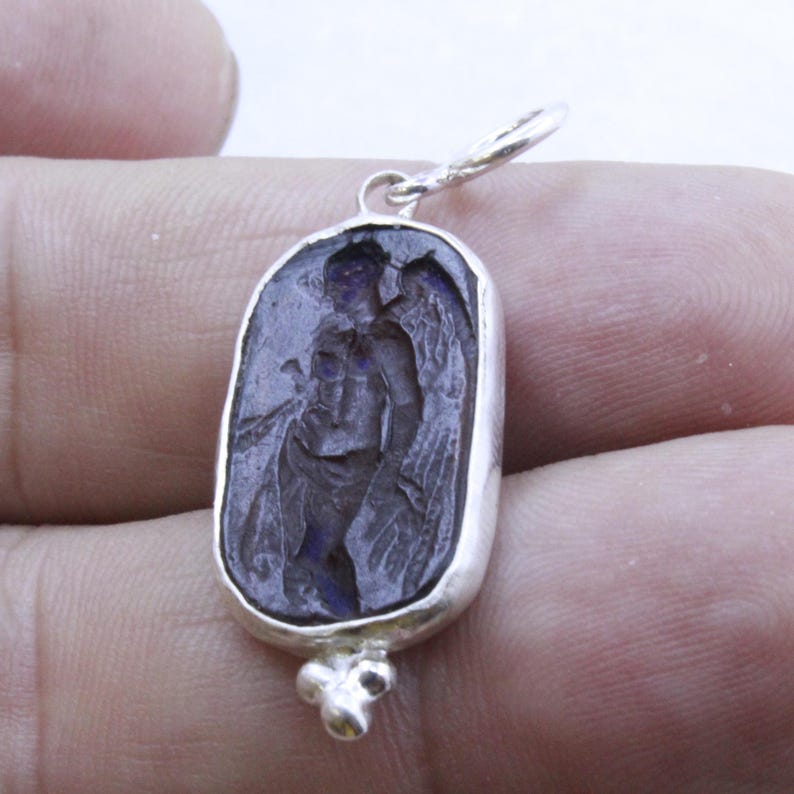 May include: A silver pendant with an oval, dark blue stone featuring an engraved image of a winged figure. The figure is holding an object, and the pendant has a small loop at the top for a chain and a decorative cluster at the bottom.