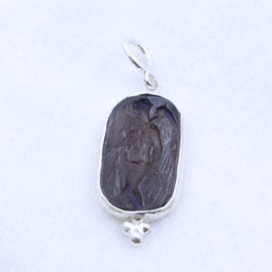 May include: A silver pendant with a carved, dark purple figure of a winged person. The pendant has a rectangular shape with rounded edges and a small decorative cluster at the bottom. The figure is detailed with visible wings and body features.