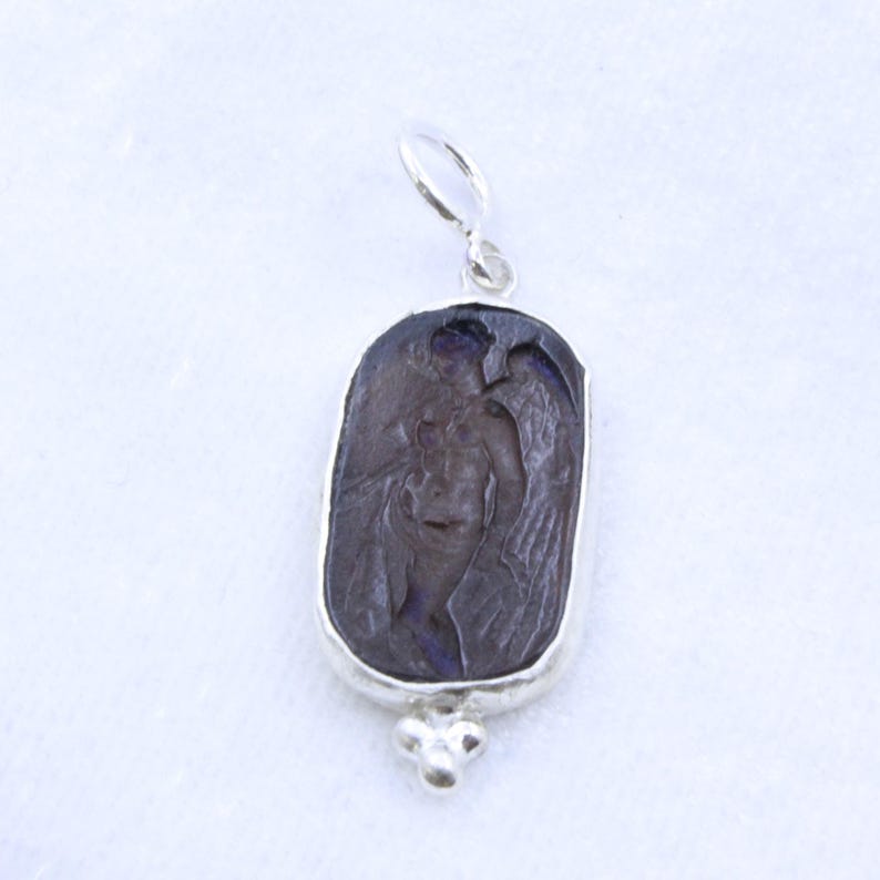 May include: A silver pendant with a carved, dark purple figure of a winged person. The pendant has a rectangular shape with rounded edges and a small decorative cluster at the bottom. The figure is detailed with visible wings and body features.